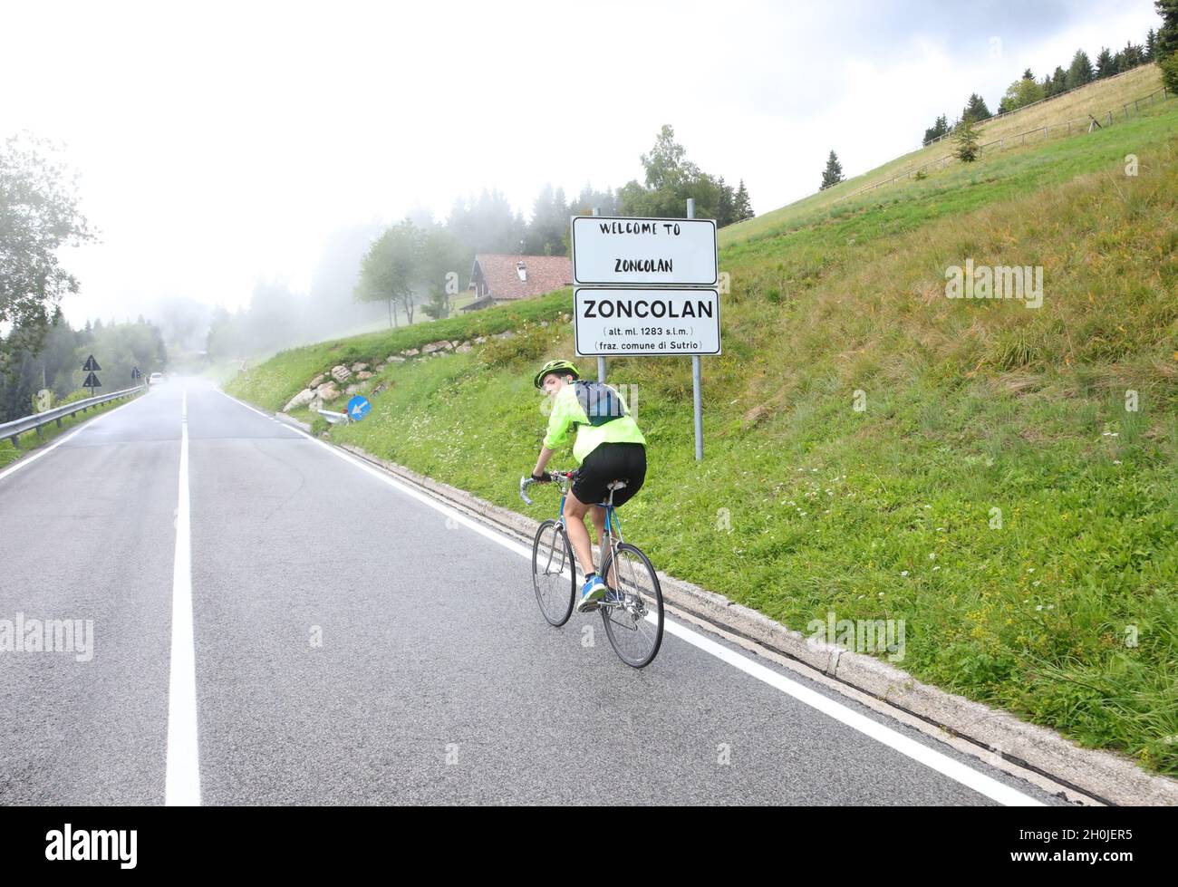 Junger Radfahrer mit Schutzhelm, der an einem regnerischen Tag mühsam auf der bergauf führenden Straße nach Monte Zoncolan in Italien mit Regenkleidung tritt Stockfoto