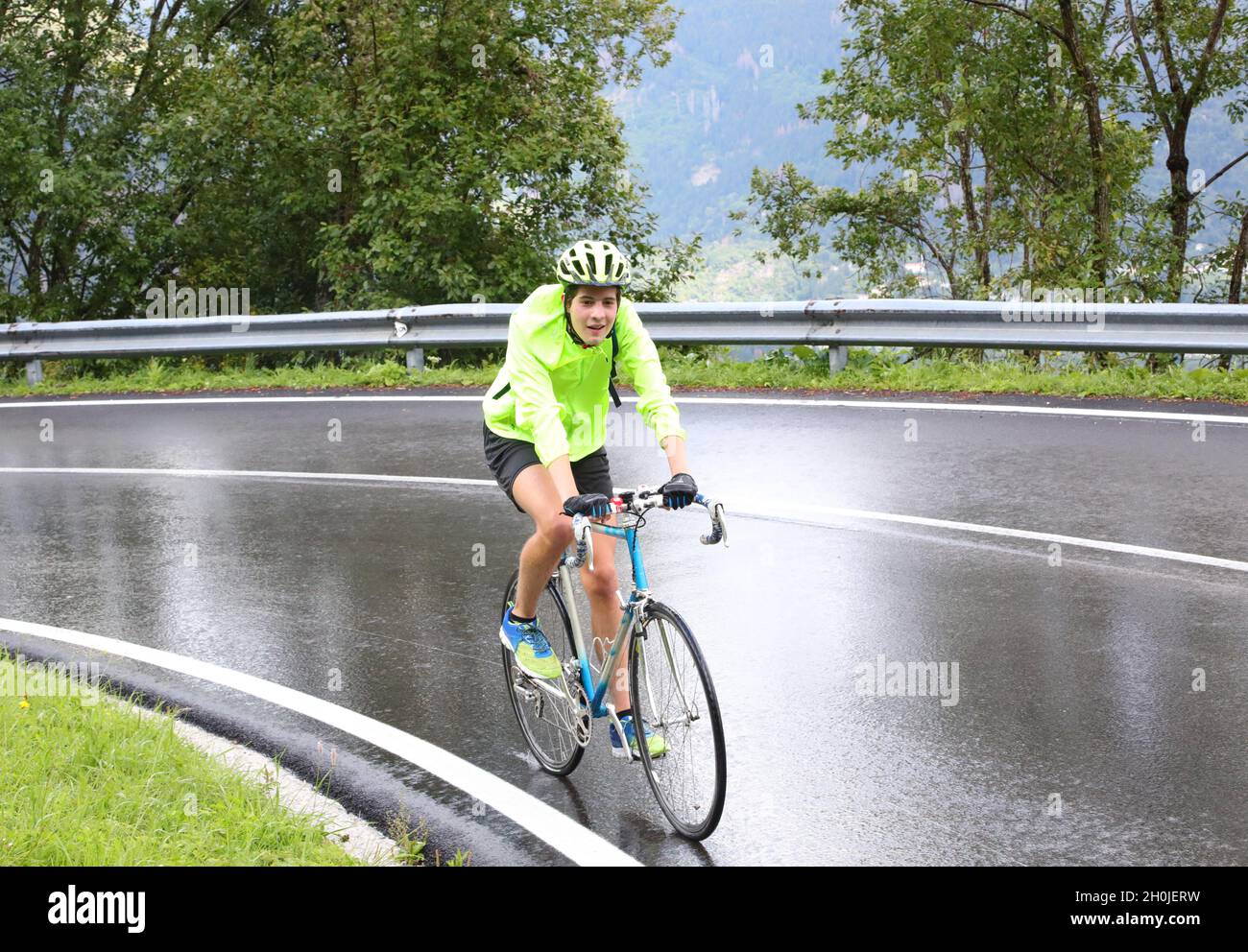 Junger Radfahrer mit Schutzhelm, der an einem regnerischen Tag mühsam mit Regenkleidung auf die Straße tritt Stockfoto