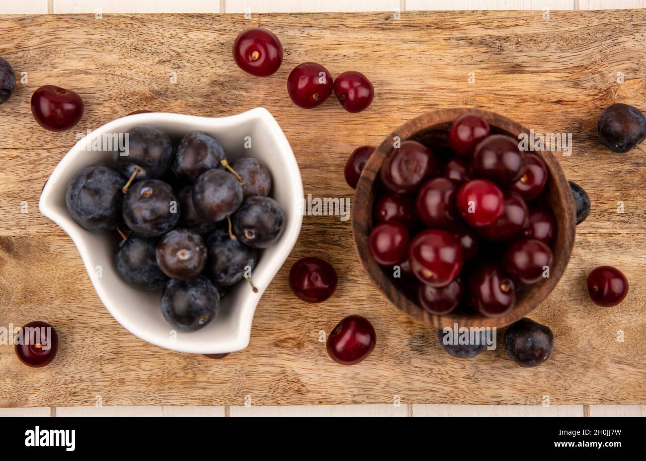 Draufsicht auf die kleinen sauer-blau-schwarzen Obstschwalben auf einer weißen Schüssel mit roten Kirschen auf einer Holzschüssel auf einem hölzernen Küchentafel auf weißem Hintergrund Stockfoto