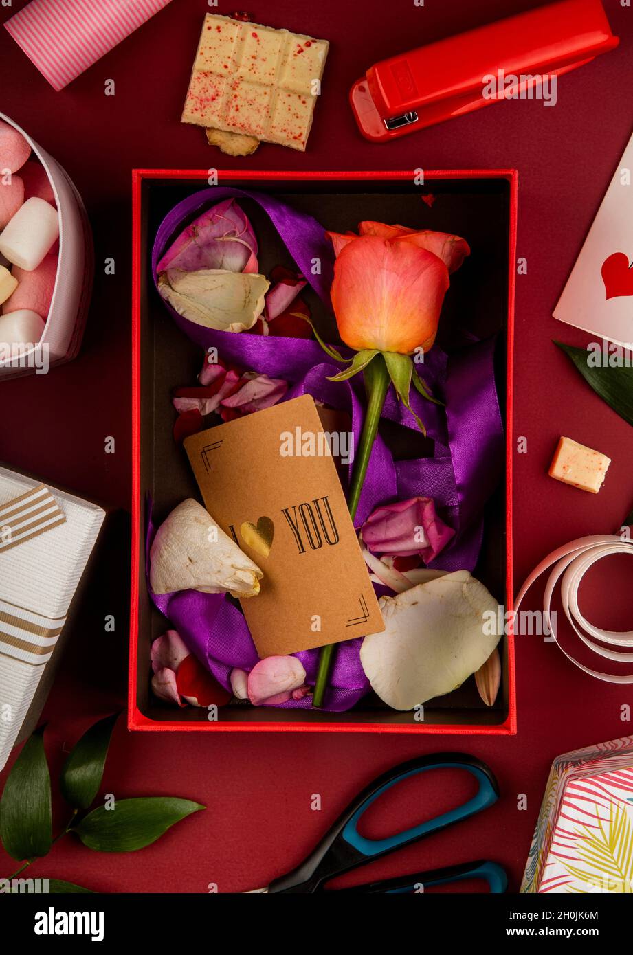 Draufsicht auf eine offene Geschenkbox mit korallenfarbener Rosenblüte und kleiner Papierkarte mit violettem Band und Blütenblättern auf rotem Hintergrund mit Schere, Hefter, Stockfoto