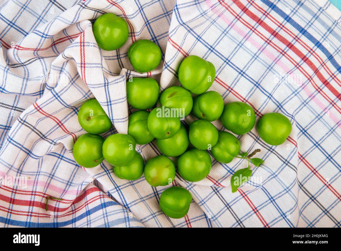 Draufsicht auf saure grüne Pflaumen, die auf karierten Stoffhintergrund verstreut sind Stockfoto