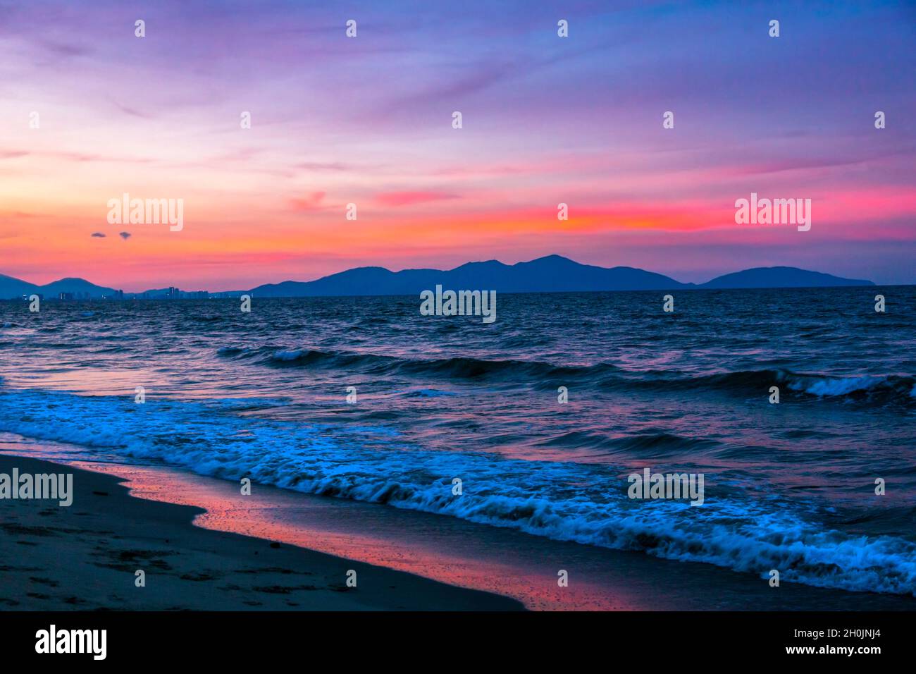 Wunderschöner Sonnenuntergang am Strand mit Rot-, Blau- und Magentaroten, mit dem Ozean und den Bergen in der Szene. Stockfoto