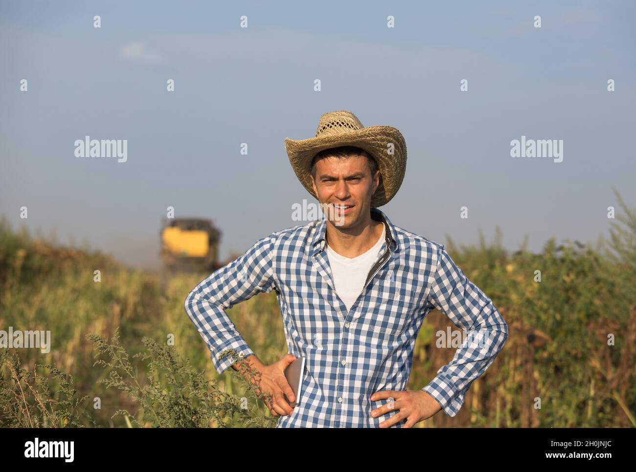 Hübscher Bauer mit Strohhut, der die Tablette hält und vor dem Mähdrescher im Sojabohnenfeld steht Stockfoto
