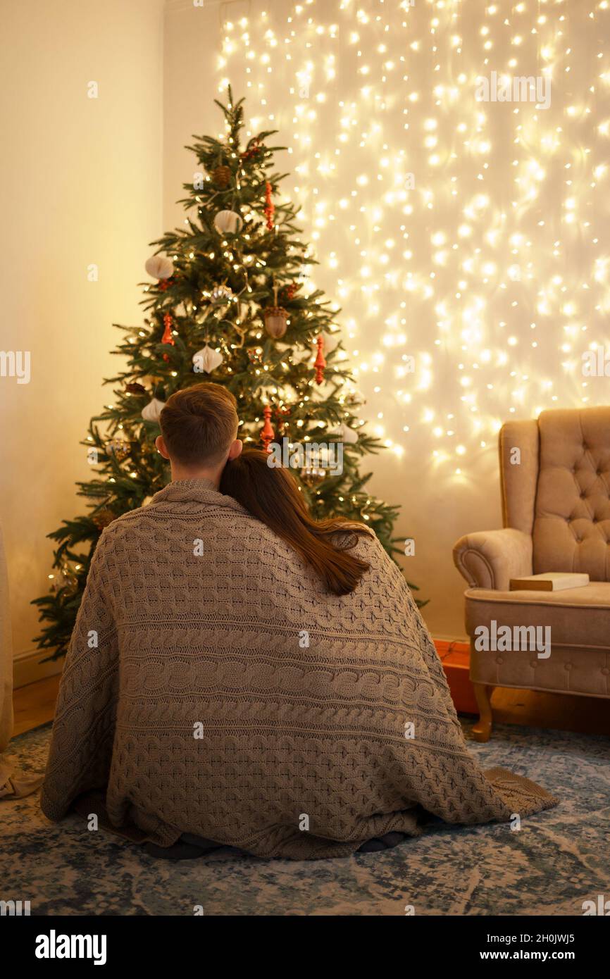 Glückliches Paar Rückansicht eingewickelt in eine bequeme Decke Blick auf geschmückten Weihnachtsbaum, während auf dem Boden zu Hause sitzen Stockfoto