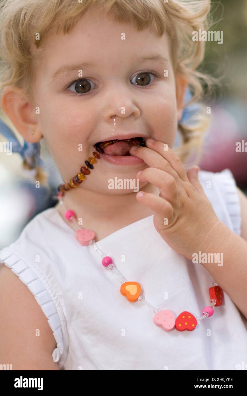 Kleines Mädchen mit laufender Nase, das eine bernsteinfarbene Halskette in den Mund legt Stockfoto