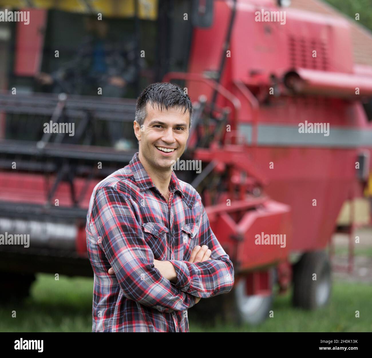 Bauer im karierten Hemd mit gekreuzten Armen, das vor dem Mähdrescher auf dem Bauernhof steht Stockfoto