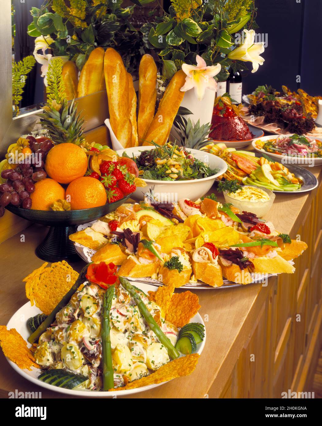 Hochformat, Buffetgerichte mit Spiegel, Blumen, Baguettes, französisches Brot, Kartoffelsalat, Geräucherter Lachs, Hühnergerichte auf einem hellen Holztisch Stockfoto