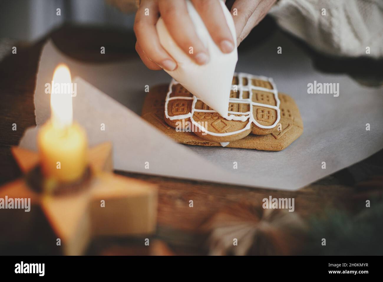 Hände schmücken weihnachts-Lebkuchenkekse mit Glasur auf Holztisch mit Kerze und Ornamenten. Nahaufnahme der Herstellung von Lebkuchenhaus mit Frostin Stockfoto