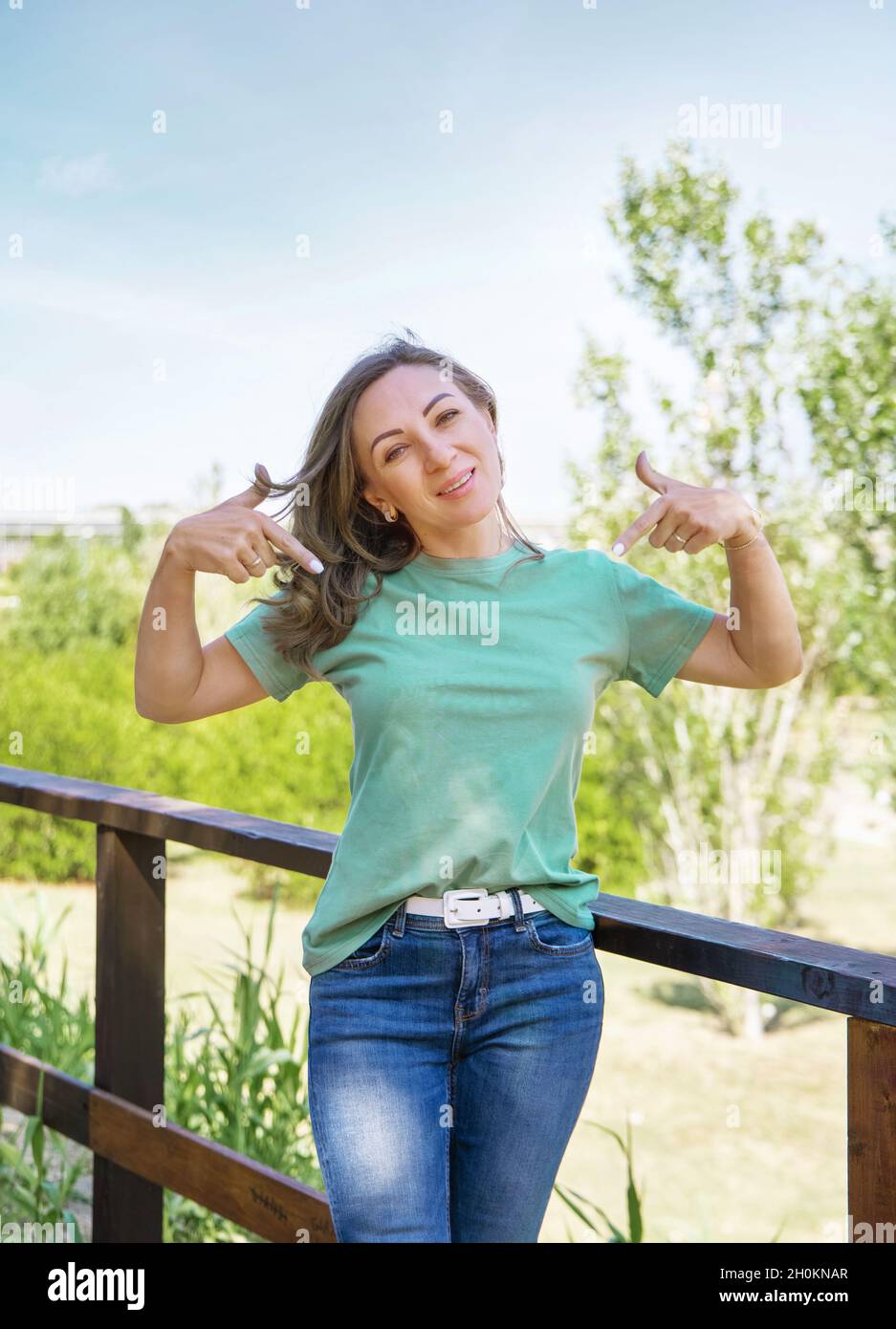 Frauen mittleren Alters, die T-Shirt und Jeans tragen, die auf T-Shirt zeigen, bleiben im Park, drei Viertel Schuss Stockfoto