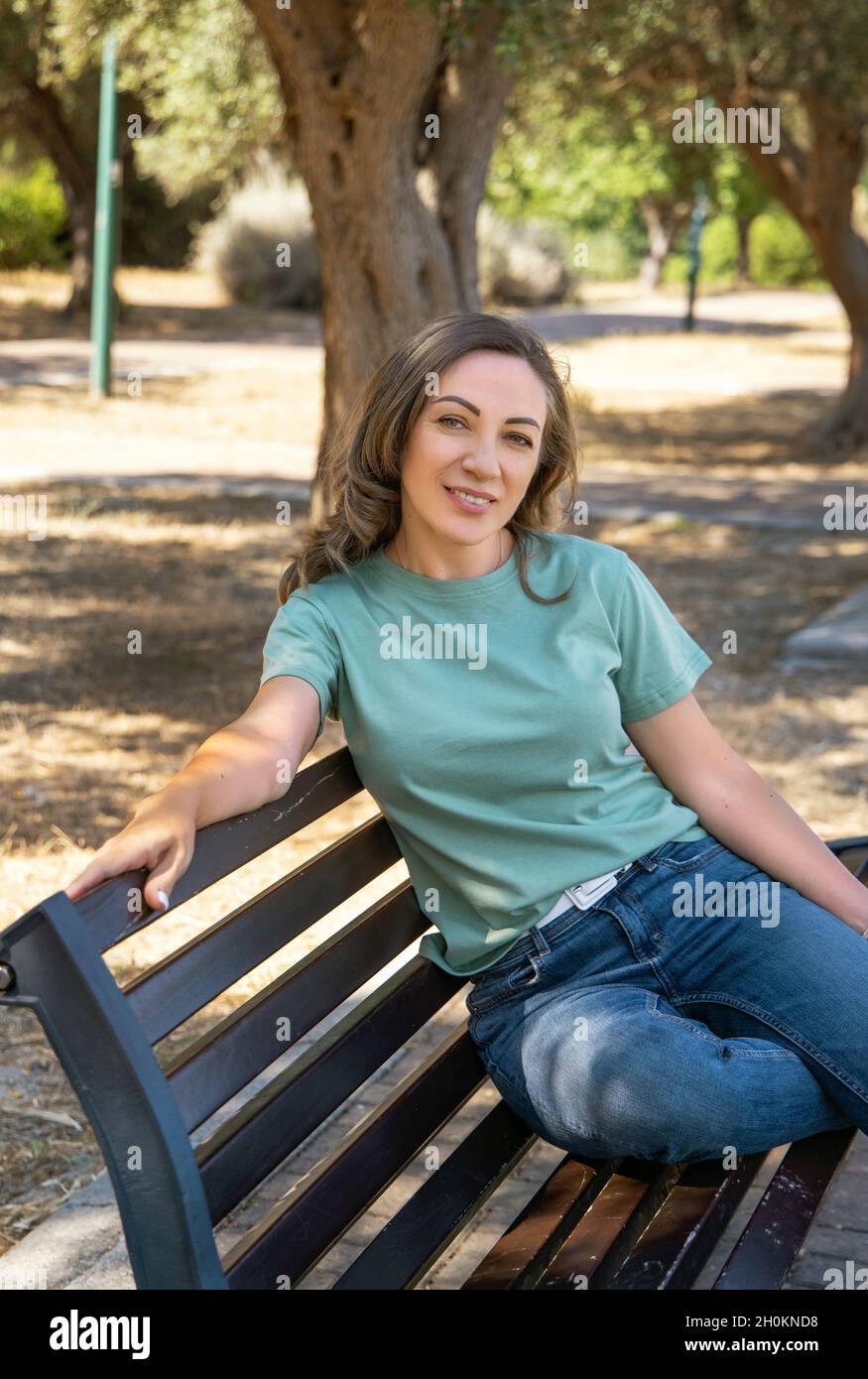 Lächelnde Frauen mittleren Alters, die T-Shirt und Jeans tragen, sitzen auf der Bank im Park und blicken auf die Kamera, Dreiviertelaufnahme Stockfoto