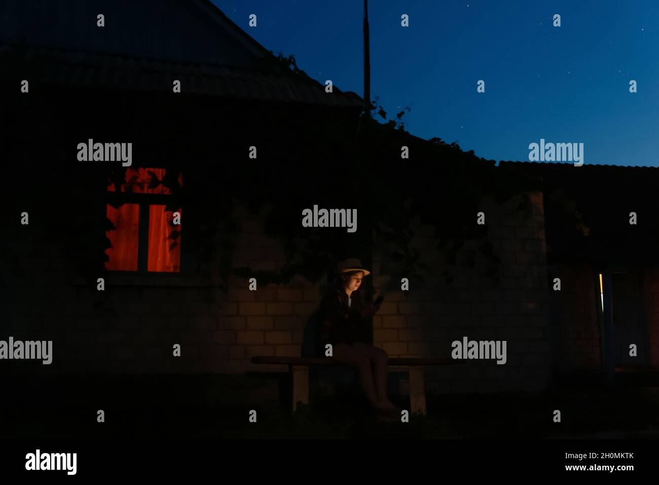 Unschärfe-Effekte für Frauen mithilfe von Smartphones, die nachts auf der Bank in der Nähe des Hauses sitzen. Außenportrait von Millennial, das nachts ihr Mobiltelefon benutzt. Orangefarbenes Licht in w Stockfoto