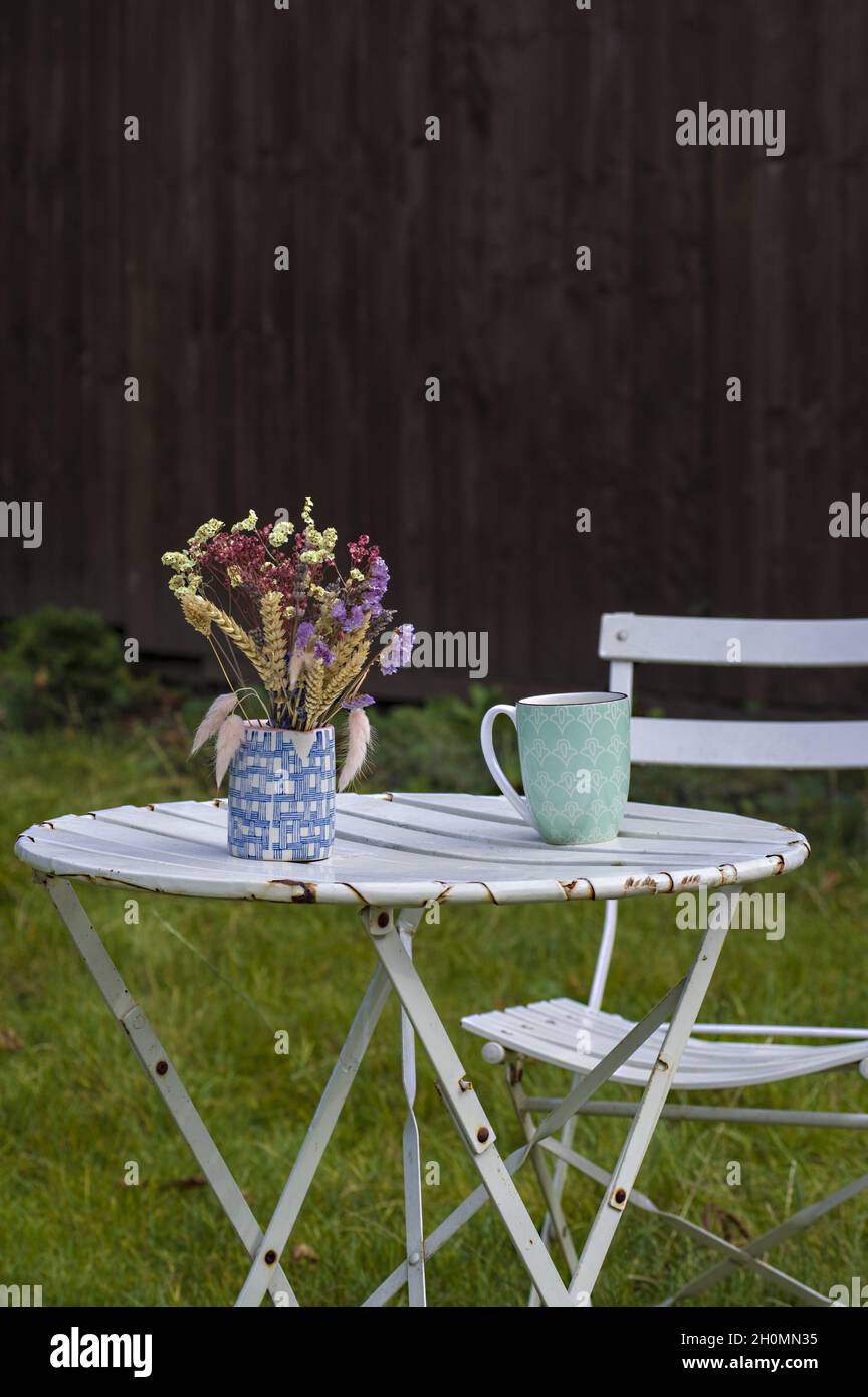 Weißer Tisch und weißer Stuhl mit einer Vase mit getrockneten Blumen und einer Teetasse oder einem Kaffeebecher draußen im Gras wie in einer Landgartenszene Stockfoto