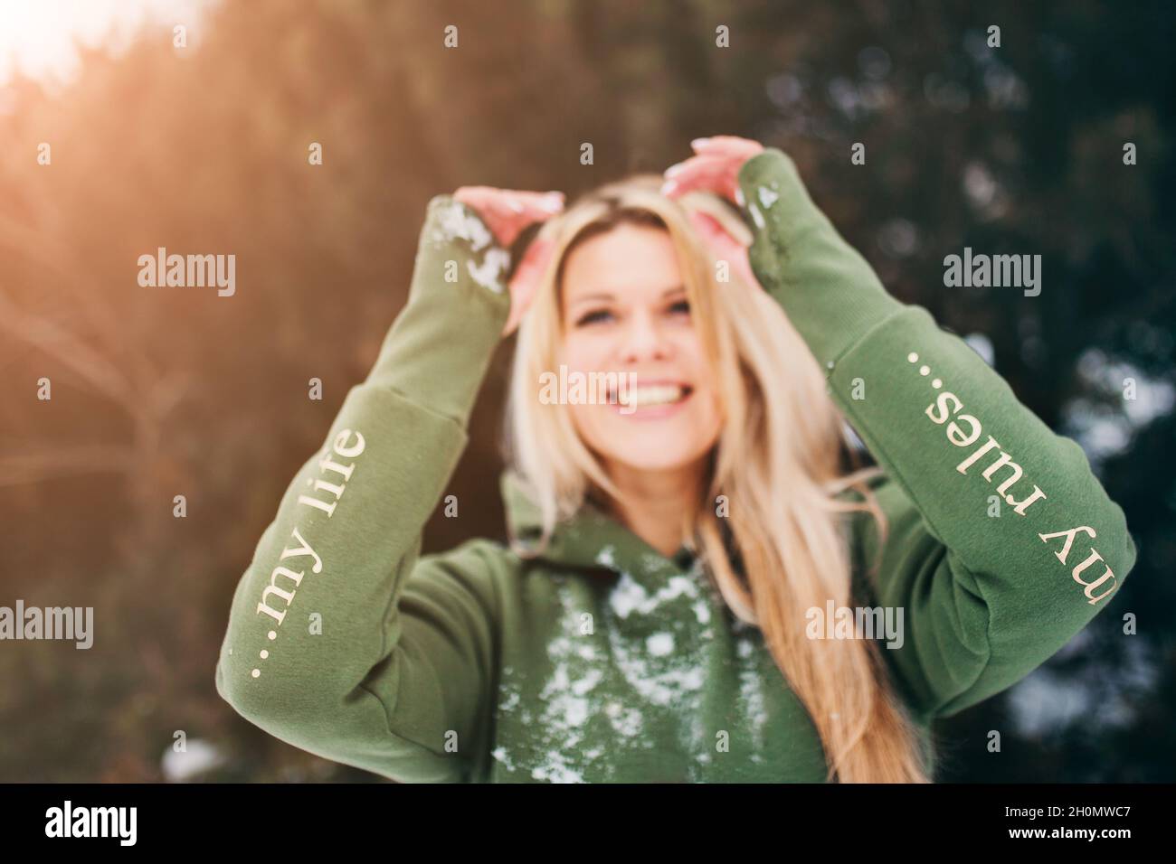 Blauäugige Blondine im grünen Hoodie im Winter im Wald - Fokus auf die Ärmel mit der Aufschrift 'Mein Leben - meine Regeln' Stockfoto