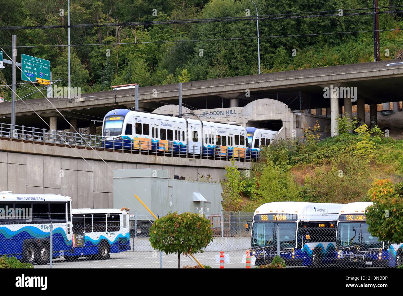 Ein Sound Transit Link-Zug fährt in den Beacon Hill Tunnel in Seattle Sodo ein, wobei die Autobahn Interstate 5 oben und ein Parkplatz für Pendlerbusse darunter liegen. Stockfoto