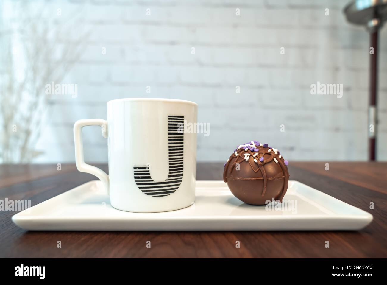 Seitenansicht einer heißen Kakao- oder Schokoladenbombe, bedeckt mit beträufelter brauner Schokolade und violetten und weißen Prise sowie einem monogrambenen J-Kaffeebecher auf der Plate Stockfoto
