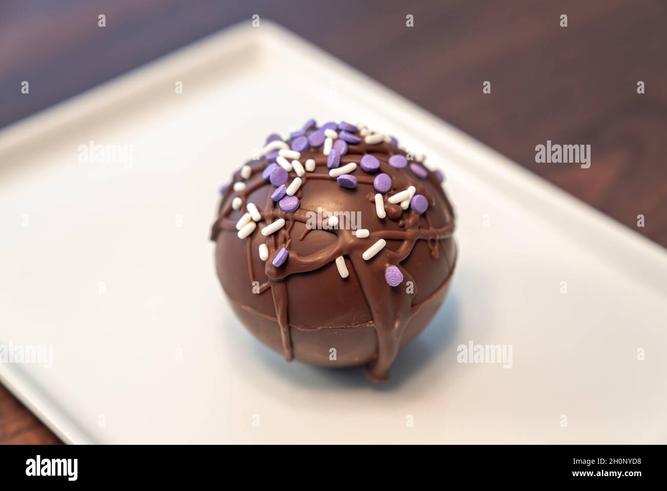 Nahaufnahme eines Lebensmittelfotos einer heißen Kakao- oder Schokoladenbombe, die mit beträufiger brauner Schokolade und violetten und weißen Prise auf einem rechteckigen Grundplatt bedeckt ist Stockfoto