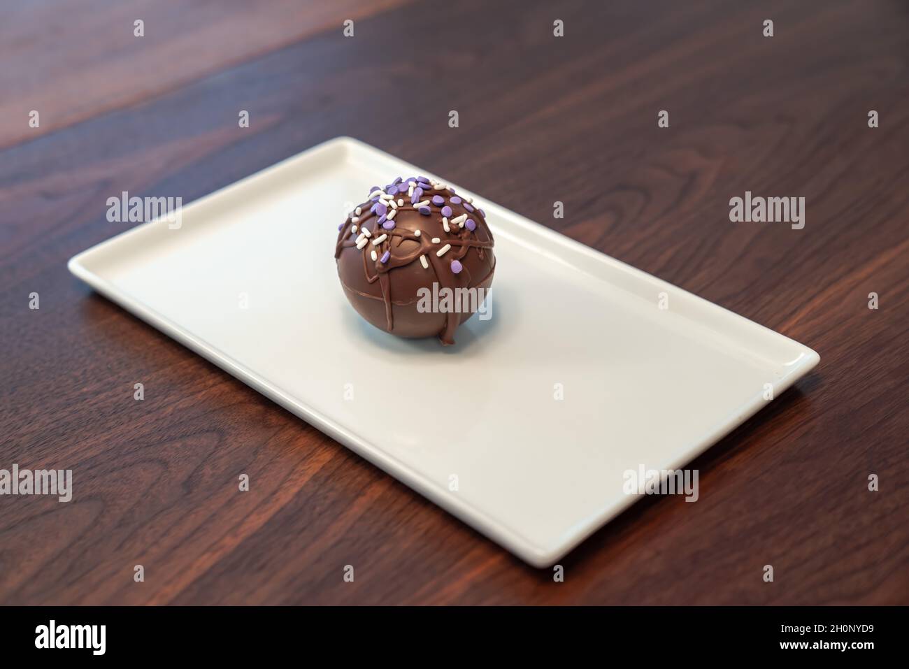 Lebensmittelfoto einer einzelnen heißen Kakao- oder Schokoladenbombe, bedeckt mit beträufelter brauner Schokolade und violetten und weißen Prise auf einem weißen Serviertafeln Stockfoto