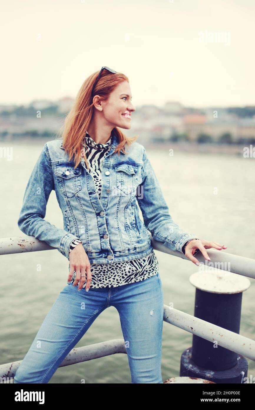 Glückliche positive Rotschopf kaukasische Frau in Denim Jacke und blaue Jeans posiert am Flussufer Stockfoto