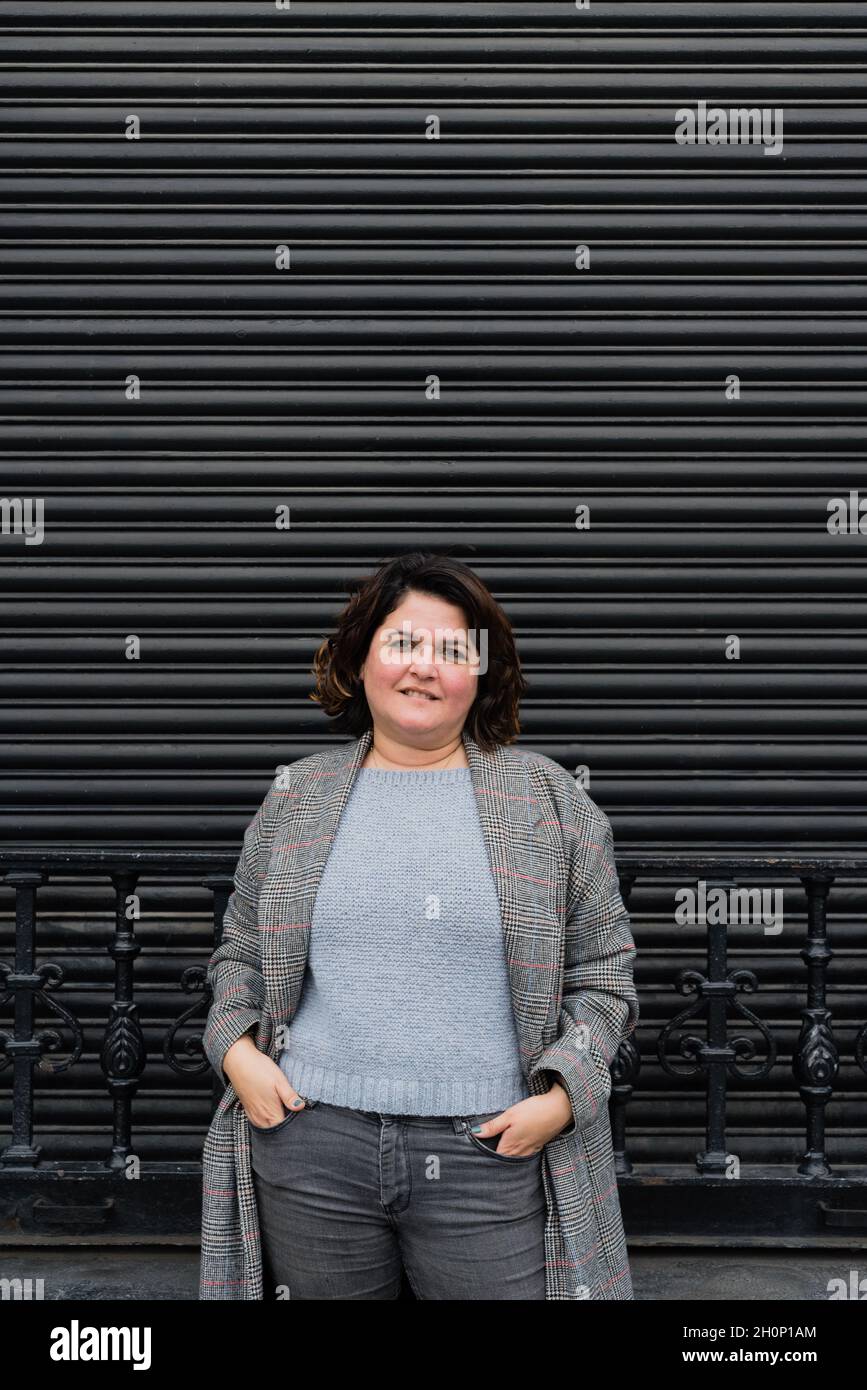 Vertikales Porträt einer Frau, die eine lange Jacke mit grauer Skala trägt, die Kamera anlächelt und ihre Hände in den Taschen hält. Speicherplatz kopieren Stockfoto