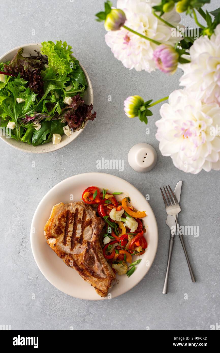 Köstliches herzhaftes Abendessen auf Tellern auf dem Tisch und ein Bouquet von schönen Herbstblumen, gebratenes Schweinefleisch-Entrecote auf dem Knochen mit geschmortem Gemüse Stockfoto