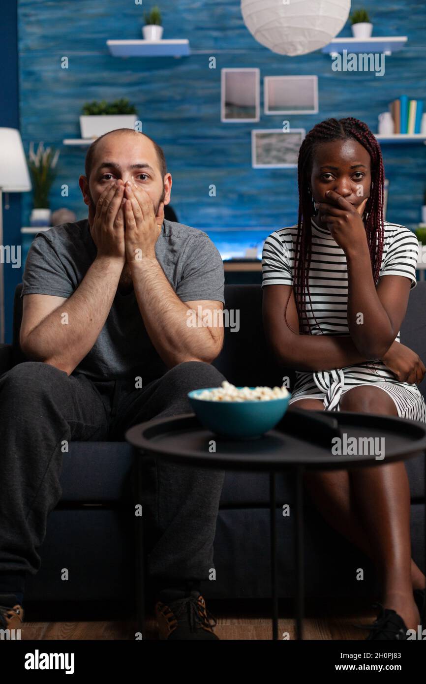 POV des interracial Paares, das schockiert ist, als es im Wohnzimmer einen Theaterfilm im Fernsehen ansieht. Gemischte Rennpartner mit den Händen über dem Mund und Blick auf Kamera und Fernsehen. Multiethnische Liebhaber Stockfoto