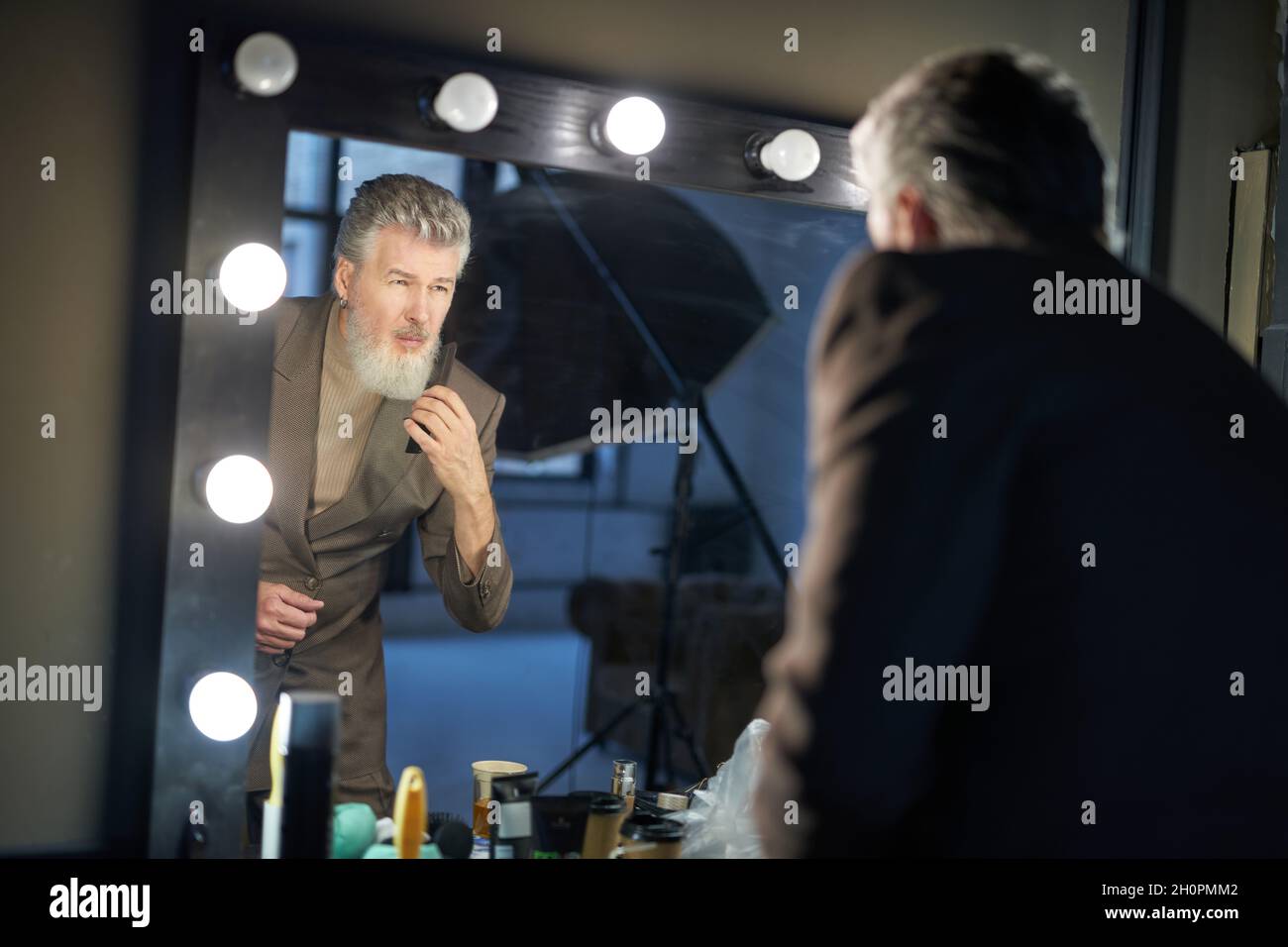 Hübscher grauhaariger Mann mittleren Alters, der einen eleganten Anzug trägt und sich im Spiegel ansieht, während er sich auf das Fotoshooting im Studio vorbereitet Stockfoto