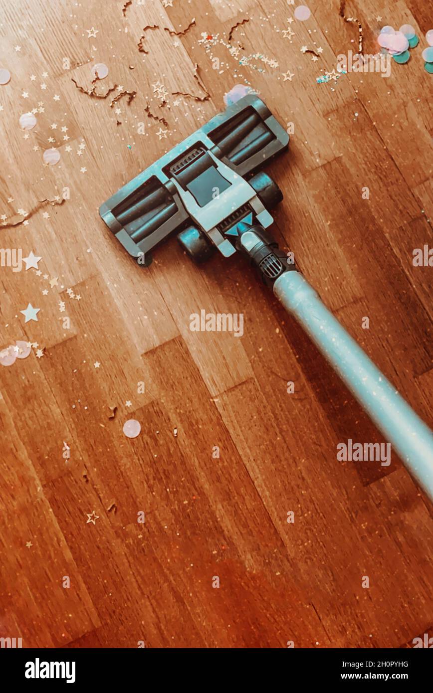Reinigung Wohnung nach den Weihnachtsferien. Ausgeschnittene Aufnahme der Staubsaugerdüse auf Holzboden-Parkett mit Konfetti zu Hause, vertikales Foto Stockfoto