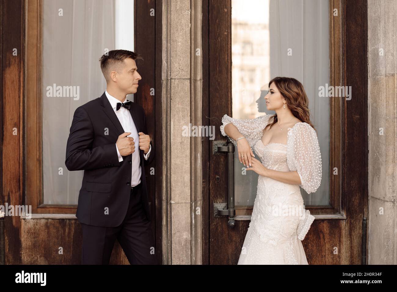 Foto von Braut und Bräutigam, die miteinander reden und sich vor dem Hintergrund der Türen anschauen. Eingang zum Gebäude. Hochzeitskleid, sui Stockfoto