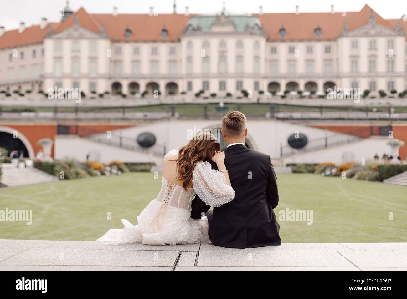 Hochzeitstag, Foto von Braut und Bräutigam, Rückansicht in Umarmung sitzend und Blick auf das Gebäude der alten Stadt im europäischen Stil. Stockfoto
