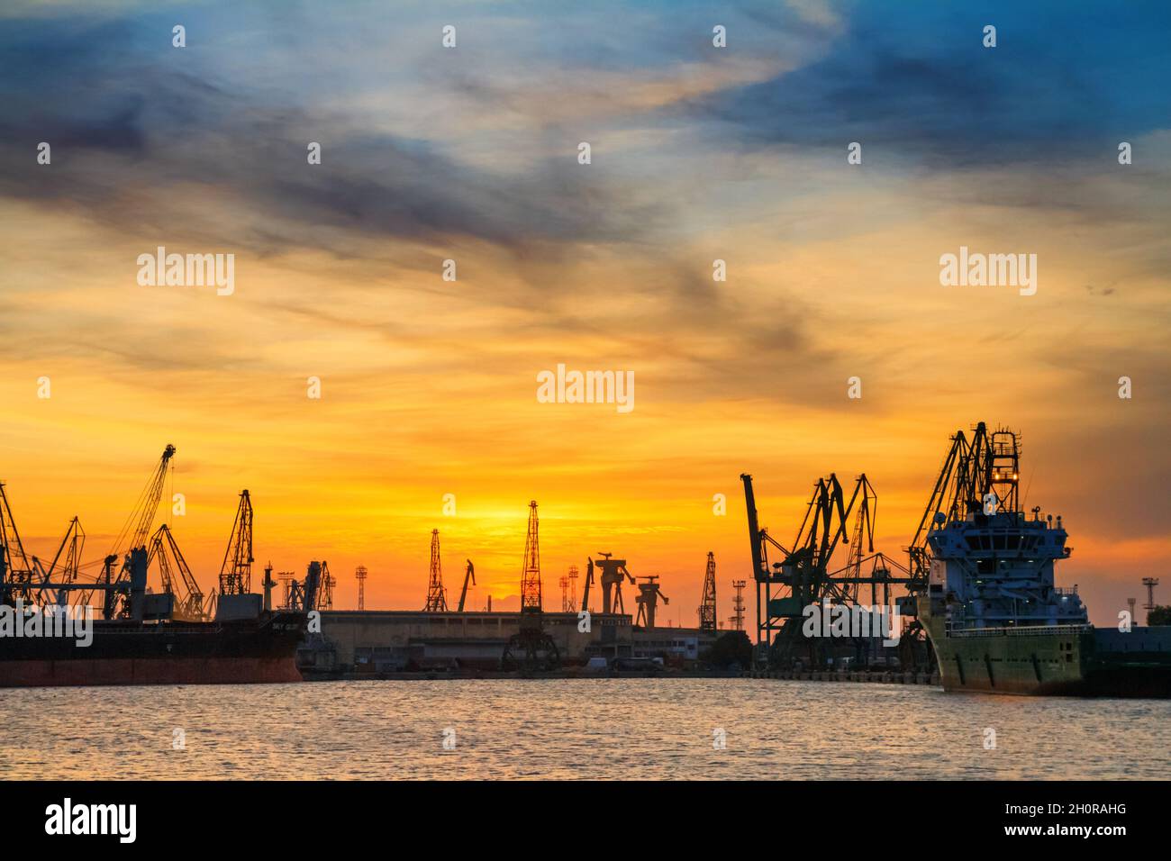 Silhouetten von Hafenkranen und Frachtschiffen im Seehafen vor dem Hintergrund des Sonnenuntergangs in Varna an der bulgarischen Schwarzmeerküste Stockfoto