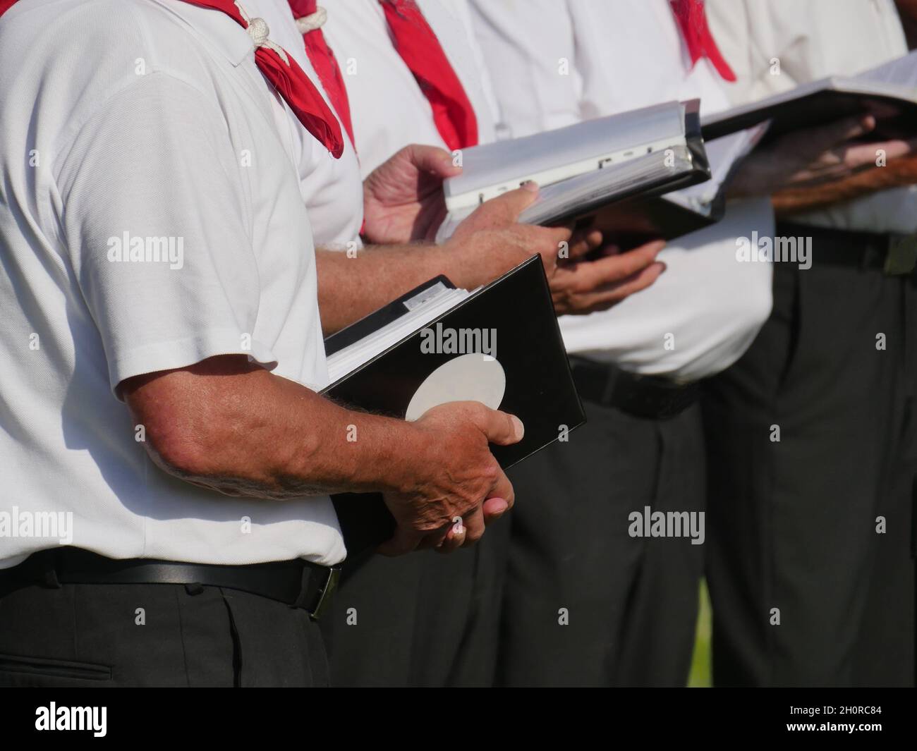 Teilansicht von 3 Sängern eines Chores, die Lehrbücher in der Hand halten, dunkle Hosen, weißes Kurzarm-Hemd, rotes Tuch, verschwommener Hintergrund Stockfoto