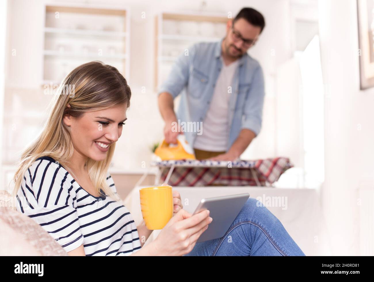Schöne junge Frau, die sich auf dem Sofa ausruht und auf das Tablet schaut, während der Mann ihr Hemd im Hintergrund bügelt. Ehemann macht Aufgaben Konzept Stockfoto