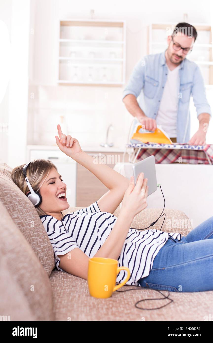 Schöne junge Frau, die sich auf dem Sofa ausruht und Musik auf Kopfhörern hört, während der Mann ihr Hemd im Hintergrund bügelt. Ehemann macht Aufgaben Konzept Stockfoto