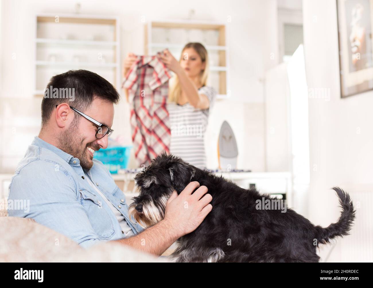Junger, hübscher Mann, der auf dem Sofa sitzt und mit dem Hund kuschelt, während die Frau im Hintergrund bügelt und Aufgaben erledigt Stockfoto
