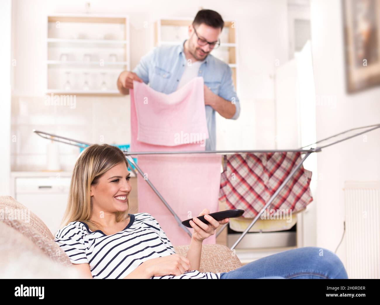 Schöne junge Frau, die sich auf dem Sofa ausruht und fernsieht, während der Mann im Hintergrund seine Wäsche wäschete. Ehemann macht Aufgaben Konzept Stockfoto
