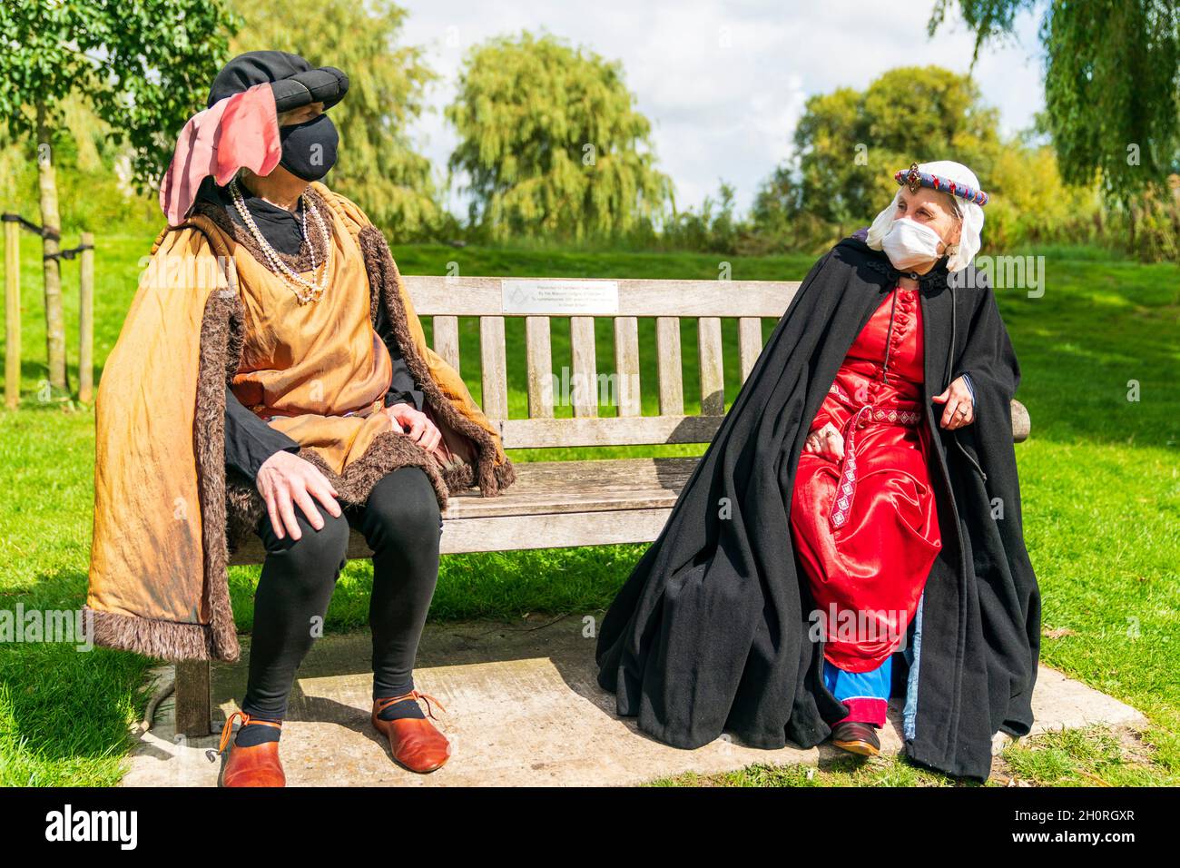 Zwei mittelalterliche Re enactors sitzen auf einer Parkbank und unterhalten sich miteinander und demonstrieren während des COVID-Sommers soziale Distanzierung und Maskentragen. Stockfoto