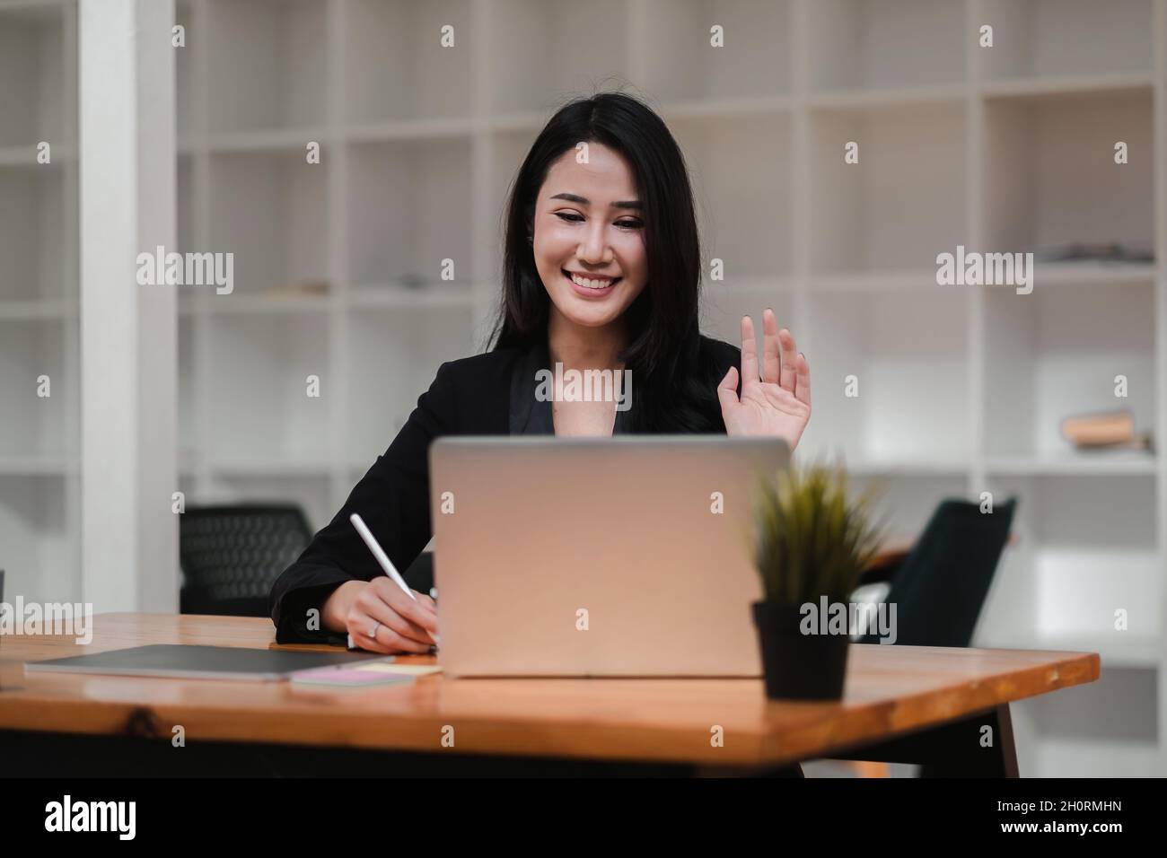 Bild einer glücklichen asiatischen Frau, die lächelt und die Hand auf den Laptop schwenkt, während sie im Büro spricht oder chattet Stockfoto
