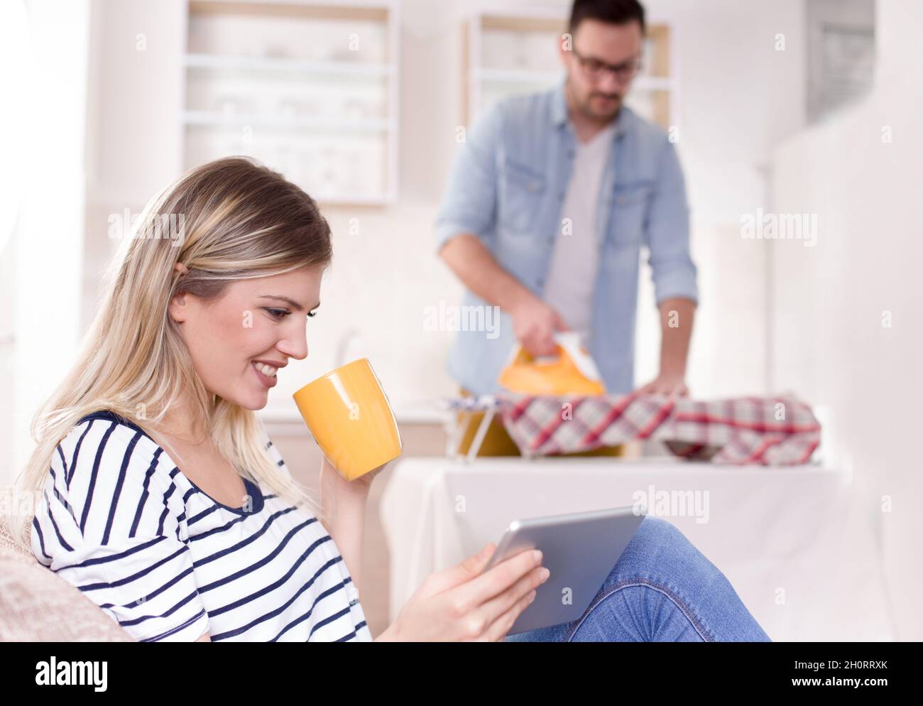 Schöne junge Frau, die sich auf dem Sofa ausruht und auf das Tablet schaut, während der Mann ihr Hemd im Hintergrund bügelt. Ehemann macht Aufgaben Konzept Stockfoto