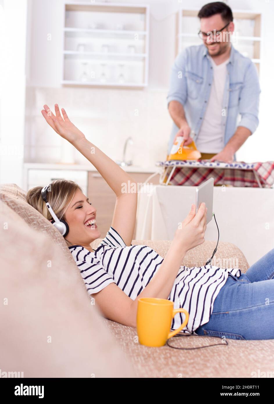 Schöne junge Frau, die sich auf dem Sofa ausruht und Musik auf Kopfhörern hört, während der Mann ihr Hemd im Hintergrund bügelt. Ehemann macht Aufgaben Konzept Stockfoto