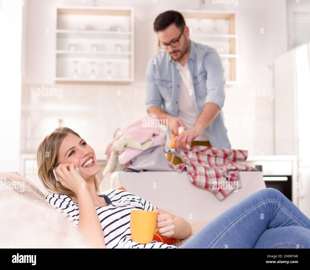 Schöne junge Frau, die sich auf dem Sofa ausruhte und auf dem Handy sprach, während der Mann ihr Hemd im Hintergrund bügelt. Ehemann macht Aufgaben Konzept Stockfoto