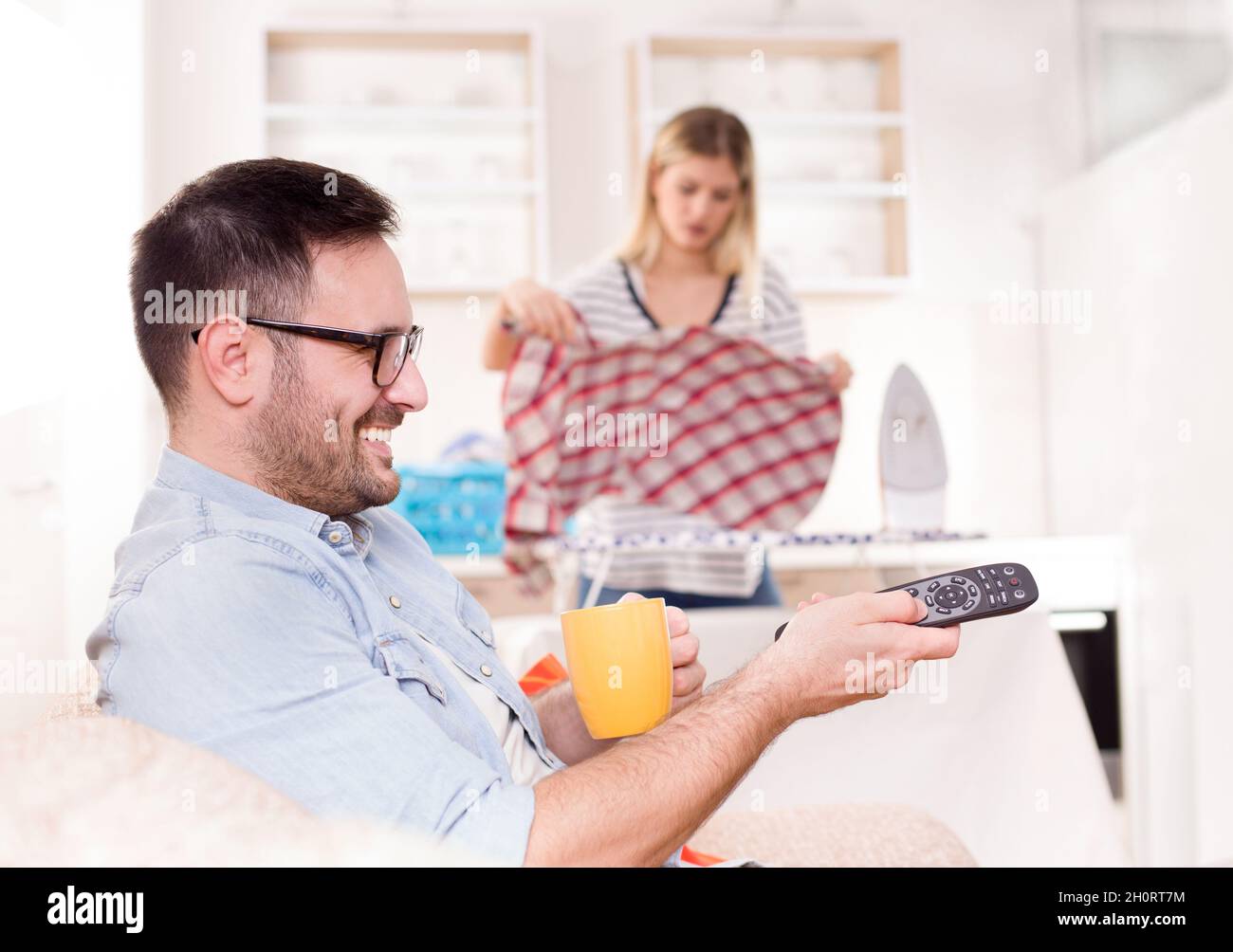 Junger, hübscher Mann, der auf dem Sofa sitzt und die Fernbedienung des fernsehers hält, während seine Frau im Hintergrund bügelt und Aufgaben erledigt Stockfoto