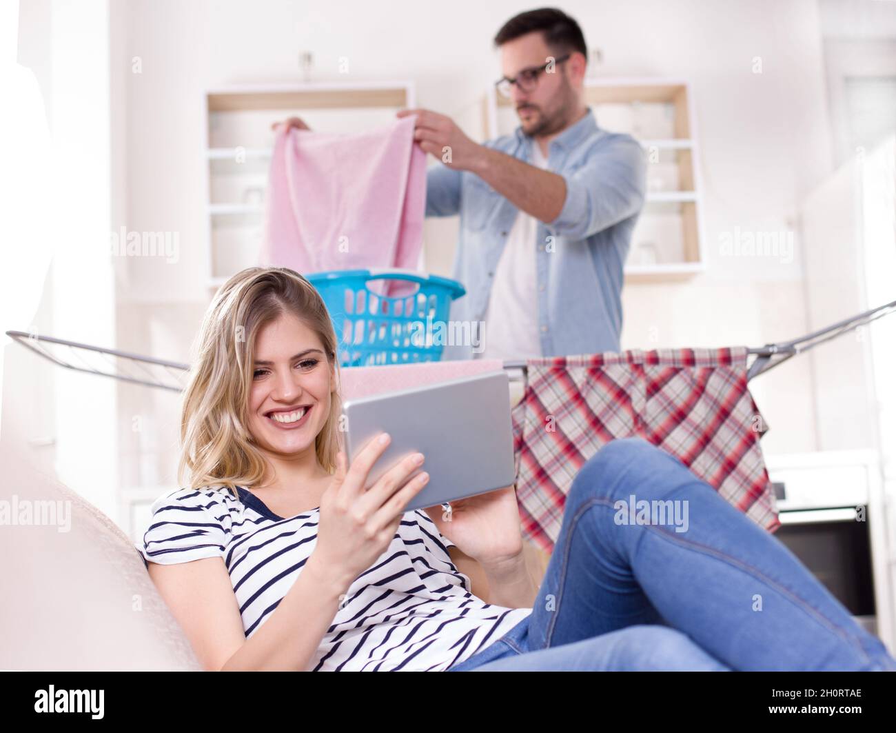 Schöne junge Frau, die sich auf dem Sofa ausruht und auf das Tablet schaut, während der Mann im Hintergrund Wäsche macht. Ehemann macht Aufgaben Konzept Stockfoto
