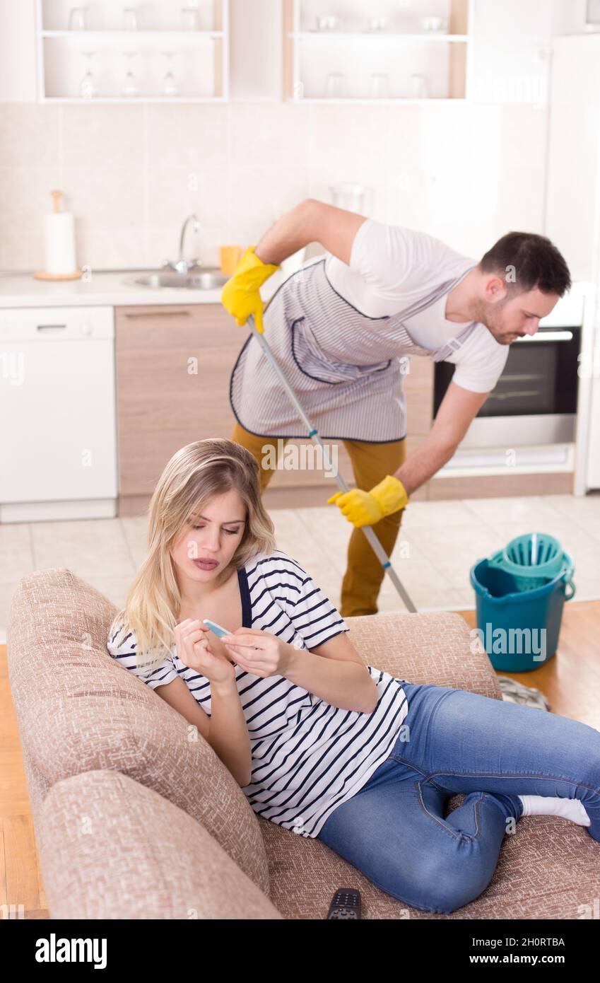 Schöne junge Frau, die sich auf dem Sofa ausruht und Maniküre macht, während der Mann im Hintergrund den Boden wischt. Ehemann macht Aufgaben Konzept Stockfoto