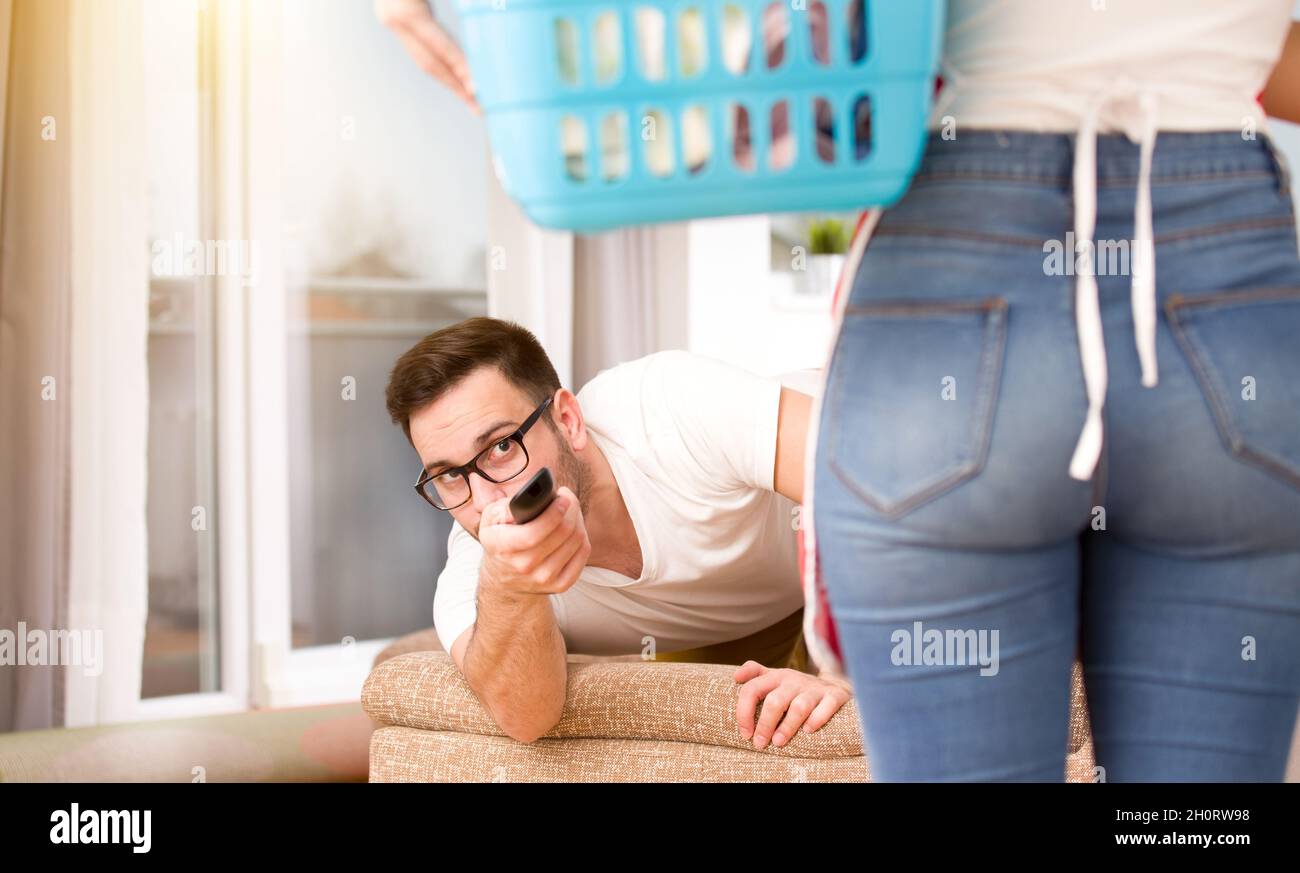 Fauler Mann mit Fernbedienung, der versucht, hinter seiner Frau mit Wäschekorb fernzusehen. Frau macht Aufgaben, während Mann sich ausruht Stockfoto