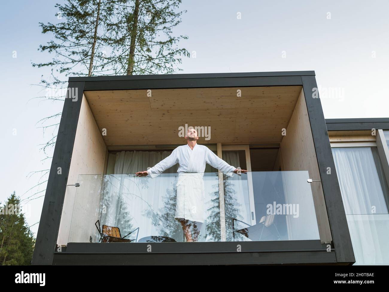 Ruhender Mann gekleideter weißer Bademantel, der auf dem Balkon des Waldhauses steht und die frische Luft mit Blick auf die Naturwälder der Pinien genießt. Außerhalb skandinavisch in Stockfoto