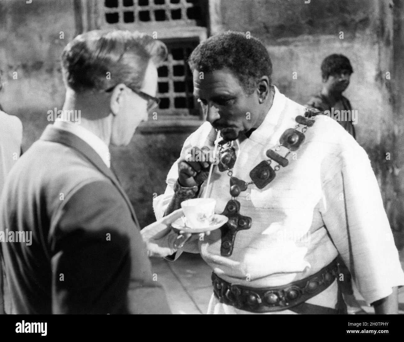 LAURENCE OLIVIER am Set trank eine Tasse Tee mit einem Stroh in Kostüm, als DER Regisseur STUART BURGE von OTHELLO 1965 William Shakespeare BHE Films / National Theatre of Great Britain Production spielt Stockfoto