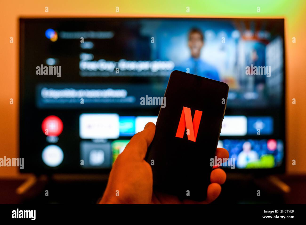 Nahaufnahme der Hand mit Smartphone und Netflix ​logo auf dem Display. Fernseher im Hintergrund. Neue Technologie, Serie, Netzwerk, Film, Kino, Comedy, e Stockfoto