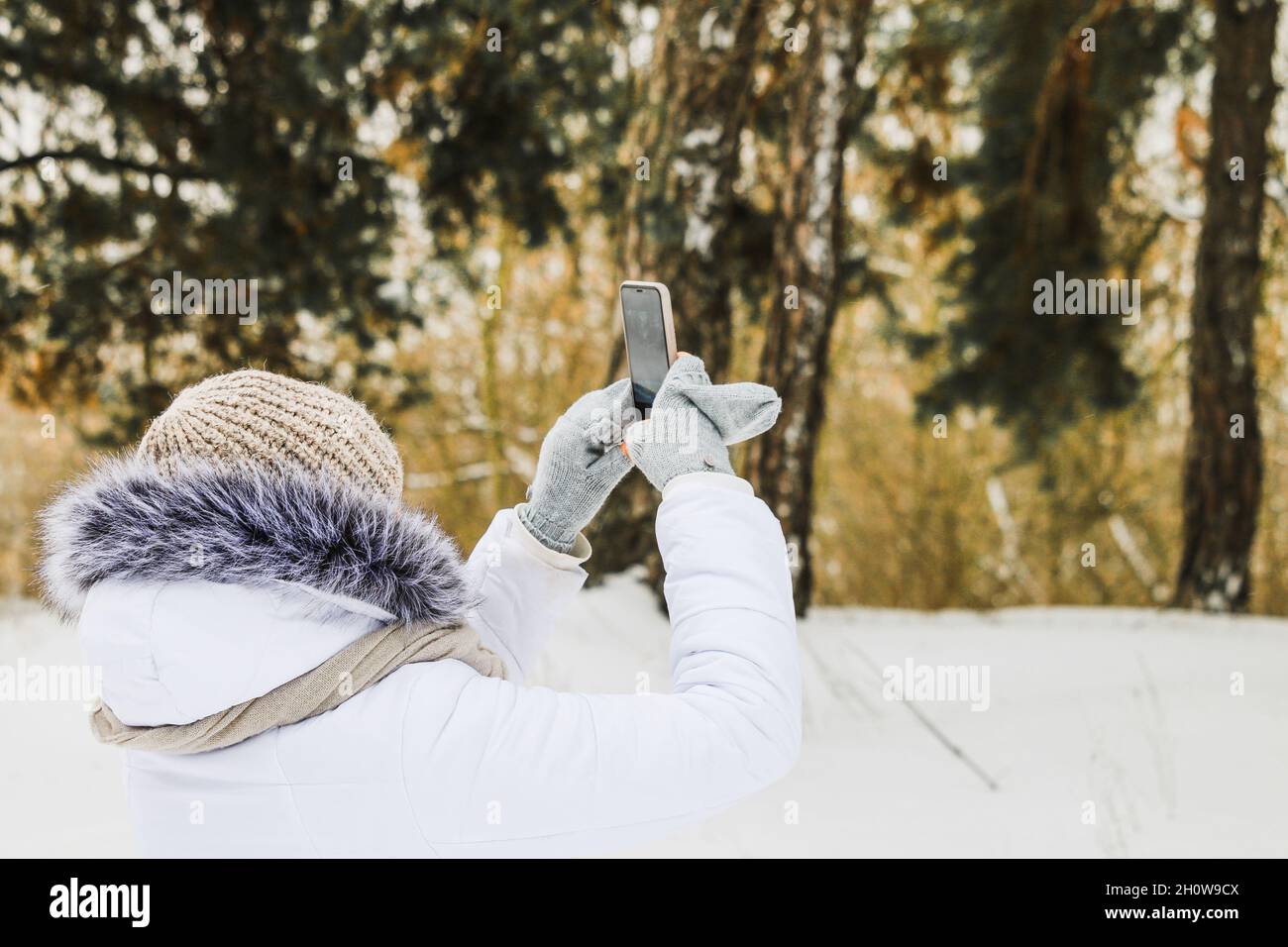 Mädchen in Weiß auf einem Spaziergang im Winterpark macht Bilder von Winterbäumen. Stockfoto