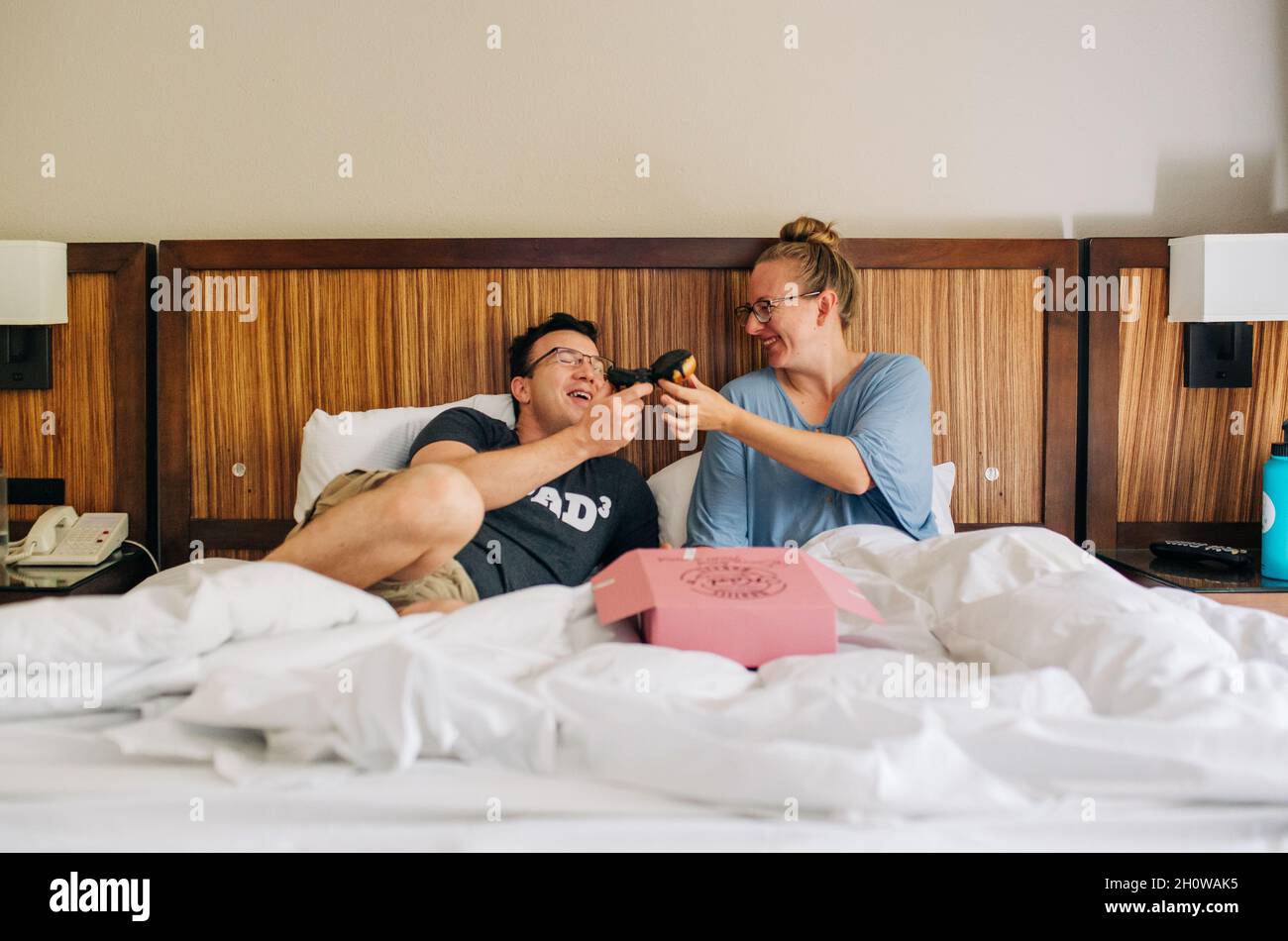 Eltern essen Donuts im Bett, während sie ihren Aufenthalt im Hotel genießen Stockfoto