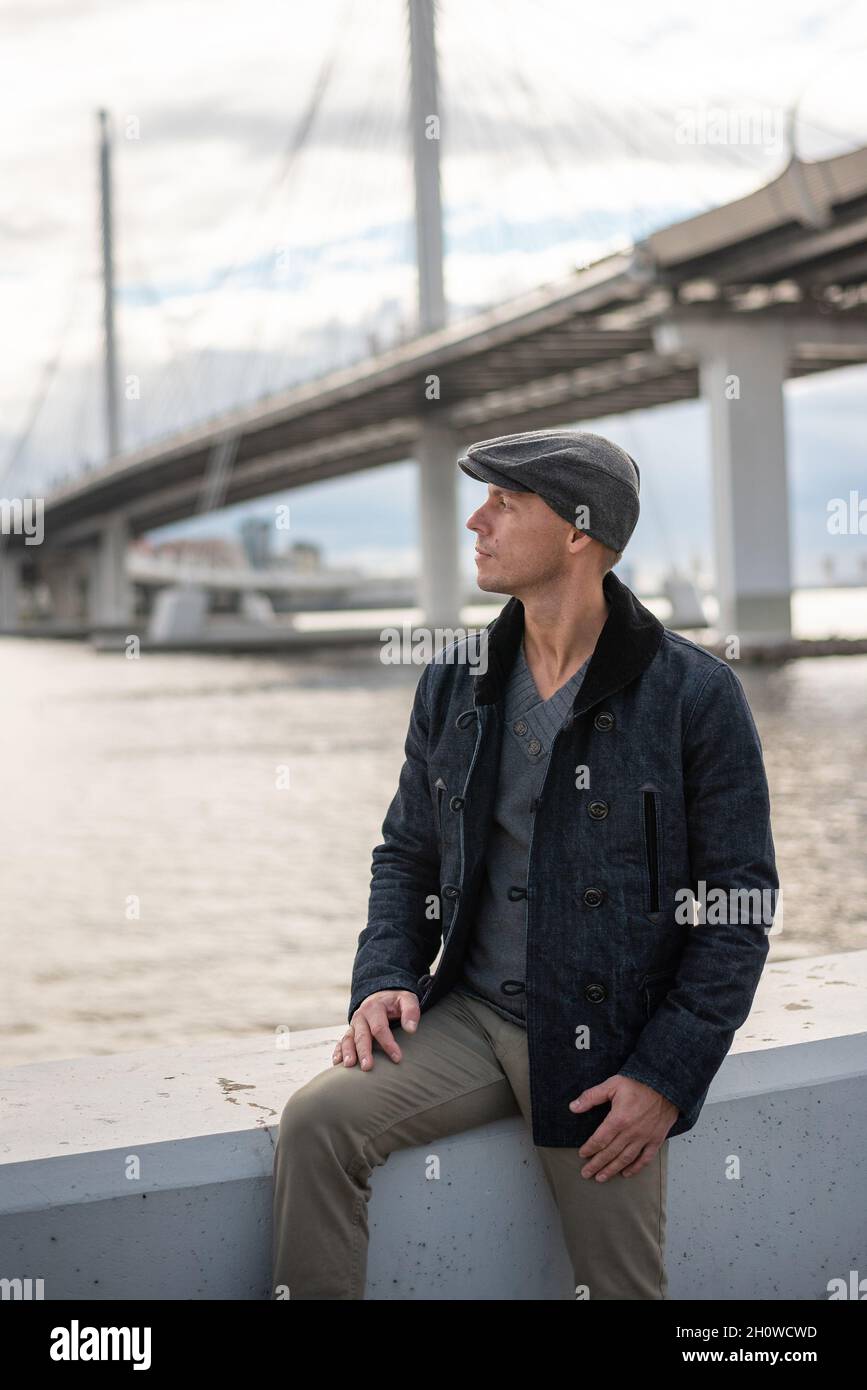 Ein Mann in einem Mantel und einer Mütze auf dem Hintergrund der Brücke. Stockfoto